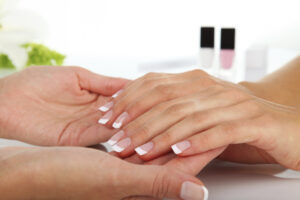 A person's hands are being held during a beauty treatment, featuring a finished French manicure. Two nail polish bottles appear out of focus in the background.